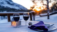 Romantic winter dinner setting with two wine glasses on snowy terrace at sunset, mountains in background, candlelight ambiance creates cozy atmosphere.