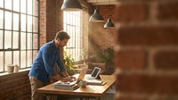 Young professional man working on laptop at wooden desk in modern loft office with brick walls and large windows during daytime.