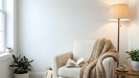 Cozy reading corner with beige armchair, knitted throw blanket, floor lamp and houseplants in bright minimalist living room interior for relaxation.