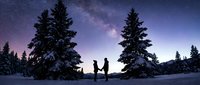 Couple standing together in snowy winter landscape under starry night sky with snow-covered evergreen trees and milky way galaxy visible overhead.