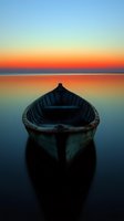 Wooden boat silhouetted against vibrant orange and blue sunset sky, reflecting on calm mirror-like water surface creating peaceful atmospheric scene.