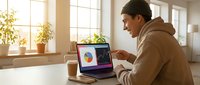 Young man of South Asian descent wearing a brown hoodie, working at a laptop displaying colorful data charts and graphs in a bright, modern office space with natural sunlight and potted plants, suitable for business, technology, and analytics content.