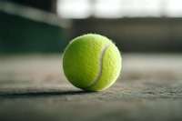 Bright yellow-green tennis ball on concrete surface with soft bokeh background, dramatic low angle perspective highlighting texture and shadows in sports equipment.