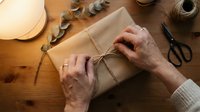 Hands wrapping kraft paper gift with twine on wooden surface, surrounded by scissors, eucalyptus branches and decorative supplies for present packaging and craft projects.