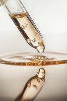 Macro shot of golden liquid with air bubbles dropping from glass pipette into liquid surface creating reflection and splash effect against light background.
