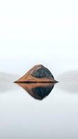 Solitary mountain peak rising from calm lake waters with perfect mirror reflection against misty background, creating symmetrical minimalist landscape composition.