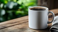 White ceramic coffee mug filled with fresh dark coffee on rustic wooden table with blurred green natural background and gray napkin, morning lifestyle.