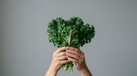Hands holding fresh kale bunch against neutral background, representing healthy eating, organic produce, nutrition, and vegetarian lifestyle choices.