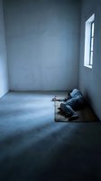 Solitary person in hooded clothing sitting alone in empty concrete room with window light creating moody atmosphere for mental health awareness concepts.
