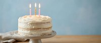 Vanilla birthday cake with white frosting and three lit candles on wooden cake stand against blue background for celebration party dessert.