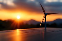 Solar panel and wind turbine silhouetted against dramatic orange sunset sky with mountain backdrop, representing renewable energy and sustainable power generation.