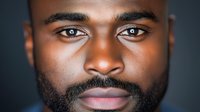 Close up portrait of young African American male with beard and intense gaze against dark background, showing confidence and determination in professional headshot style.