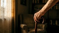 Elderly man's weathered hands gripping wooden walking cane in warm sunlit room with vintage furniture and bookshelves creating cozy atmospheric interior.