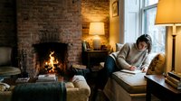 Young woman reading book in cozy living room with fireplace and warm lighting. Comfortable home interior for relaxation and leisure activities.