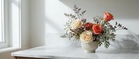 Elegant floral arrangement with orange and cream roses in a white ceramic vase on a marble surface, soft natural light, minimalist interior.