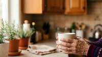 Senior woman with weathered hands holding warm beverage in cozy kitchen setting with potted plants and wooden cabinetry, lifestyle and home comfort concept.
