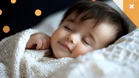 Peaceful sleeping Asian toddler wrapped in soft white blanket smiling while napping, with warm bokeh lights in background creating cozy atmosphere.