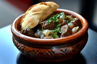 Tender beef stew with onions and fresh herbs served in traditional clay pot with crusty bread, rich brown gravy sauce in rustic ceramic bowl for restaurant menu.