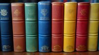 Row of vintage hardcover books with colorful spines in blue, green, red, and yellow showing decorative embossing and classic binding designs arranged on shelf.