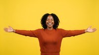 Young woman with dark curly hair and medium skin tone wearing rust-colored ribbed sweater, arms outstretched in celebratory pose against bright yellow background, expressing joy and freedom.