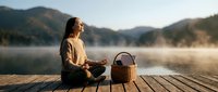 Young woman with long dark hair, wearing casual beige clothing, sitting on wooden dock at misty mountain lake at sunrise with picnic basket, peaceful outdoor leisure scene.