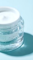 Open glass jar of white face cream with water droplets on light blue background, skincare and beauty product concept.