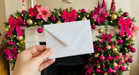 Hand holding blank white envelope against festive Christmas garland with pink ribbons and ornaments for holiday greeting cards and seasonal messaging.