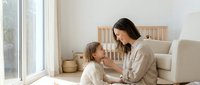 Young Caucasian mother and blonde daughter sharing tender moment on living room floor near window, bonding together in bright modern home interior.