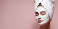 Young woman with white facial mask and towel on head enjoying spa treatment for skincare routine and relaxation on pink background.