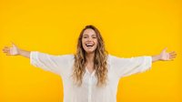 Young woman with long wavy brown hair and fair complexion wearing white button-up shirt, arms spread wide in celebratory pose against bright yellow background, expressing joy and freedom.