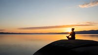 Silhouette of person meditating on rock by calm lake at golden sunset with peaceful sky reflection creating serene wellness and mindfulness atmosphere.