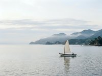 Lone sailboat gliding on calm misty waters with mountain silhouettes in background, evoking tranquility and adventure on a cloudy day.