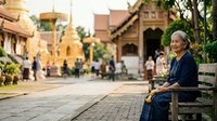 Senior woman in traditional indigo clothing sitting on bench in Southeast Asian temple courtyard with golden architecture and tourists in background.