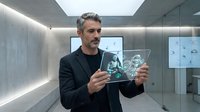 Middle-aged man with gray hair and facial hair, wearing black blazer over dark shirt, holding transparent digital tablet displaying holographic data visualization in modern minimalist corridor with concrete walls and ambient lighting.