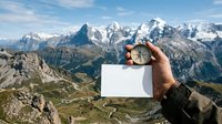 Hiker holding compass and blank white card with alpine mountain range landscape in background, navigation and outdoor adventure concept for travel and exploration themes.