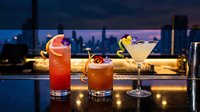 Three elegant cocktails on rooftop bar counter with city skyline at dusk. Premium drinks including martini and colorful mixed beverages for nightlife entertainment.