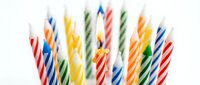Colorful striped birthday candles clustered together with one lit flame glowing brightly, isolated on a white background for celebration designs.