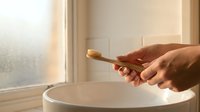 Eco-friendly bamboo toothbrush held over white ceramic sink in modern bathroom with natural lighting for sustainable oral care routine.