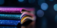Colorful glitter makeup pencils arranged in row on dark background with bokeh lights, macro photography showing sparkly cosmetic detail and texture.
