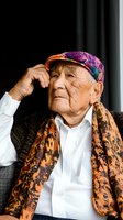Senior Asian man wearing colorful patterned cap and leopard print scarf, white shirt, thoughtful pose with hand to head, suitable for lifestyle, diversity, aging, and fashion editorial use.