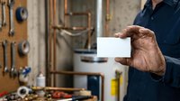 Mature male tradesman with weathered hands holding blank white card in workshop surrounded by tools and equipment for construction or repair work.