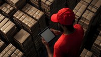 Warehouse worker in red uniform using digital tablet for inventory management among stacked cardboard boxes in modern logistics facility.