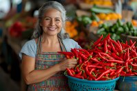 Cheerful mature Asian female vendor at farmers market displays fresh red chili peppers in blue basket, wearing striped apron and gray shirt, surrounded by produce.