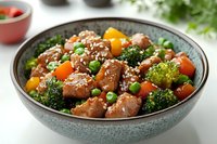 Stir-fried chicken pieces with broccoli, carrots, yellow bell peppers and green peas in teriyaki sauce, garnished with sesame seeds served in ceramic bowl.