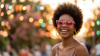 Happy young African American woman with afro hairstyle wearing pink sunglasses smiling joyfully against colorful bokeh lights background for summer lifestyle content.