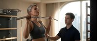 Young blonde woman doing pull-ups with personal trainer assistance in modern gym. Fitness coaching session with professional guidance for strength training workout.