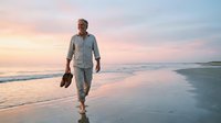 Middle-aged man with beard, wearing casual light blue shirt and jeans, walking barefoot on sandy beach at sunset holding shoes, ideal for lifestyle and travel concepts.
