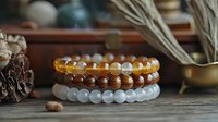 Stack of three natural stone beaded bracelets with wooden and crystal elements on rustic wooden surface with pine cone and dried plant decor in background.