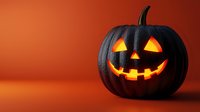 Black carved pumpkin jack-o-lantern with glowing yellow face on orange background for Halloween decorations and seasonal autumn celebrations.