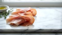 Thin slices of prosciutto ham arranged on white marble cutting board with herbs and olive oil for gourmet cooking and charcuterie preparation.